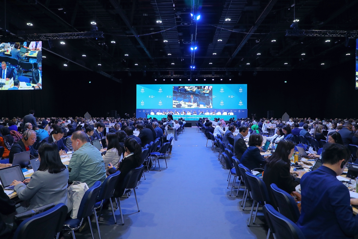 WTO Ministerial Conference. Photo of the heads of delegations meeting Abu Dhabi in a large crowded hall, looking along the central aisle to a brightly lit podium in the distance