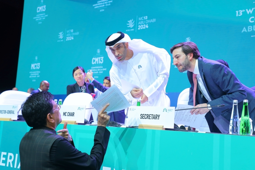 Photo of the podium at the Abu Dhabi WTO Ministerial Conference. Indian minister Puyash Goyal is in front of the podium discussing a document with conference chair Thani bin Ahmed Al Zeyoudi and senior Secretariat official Santiago Wills, above him